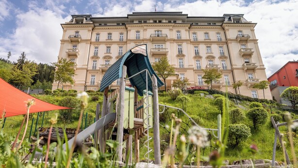 Un edificio grande de varios pisos con un estilo arquitectónico clásico, rodeado de exuberante vegetación y un parque infantil.