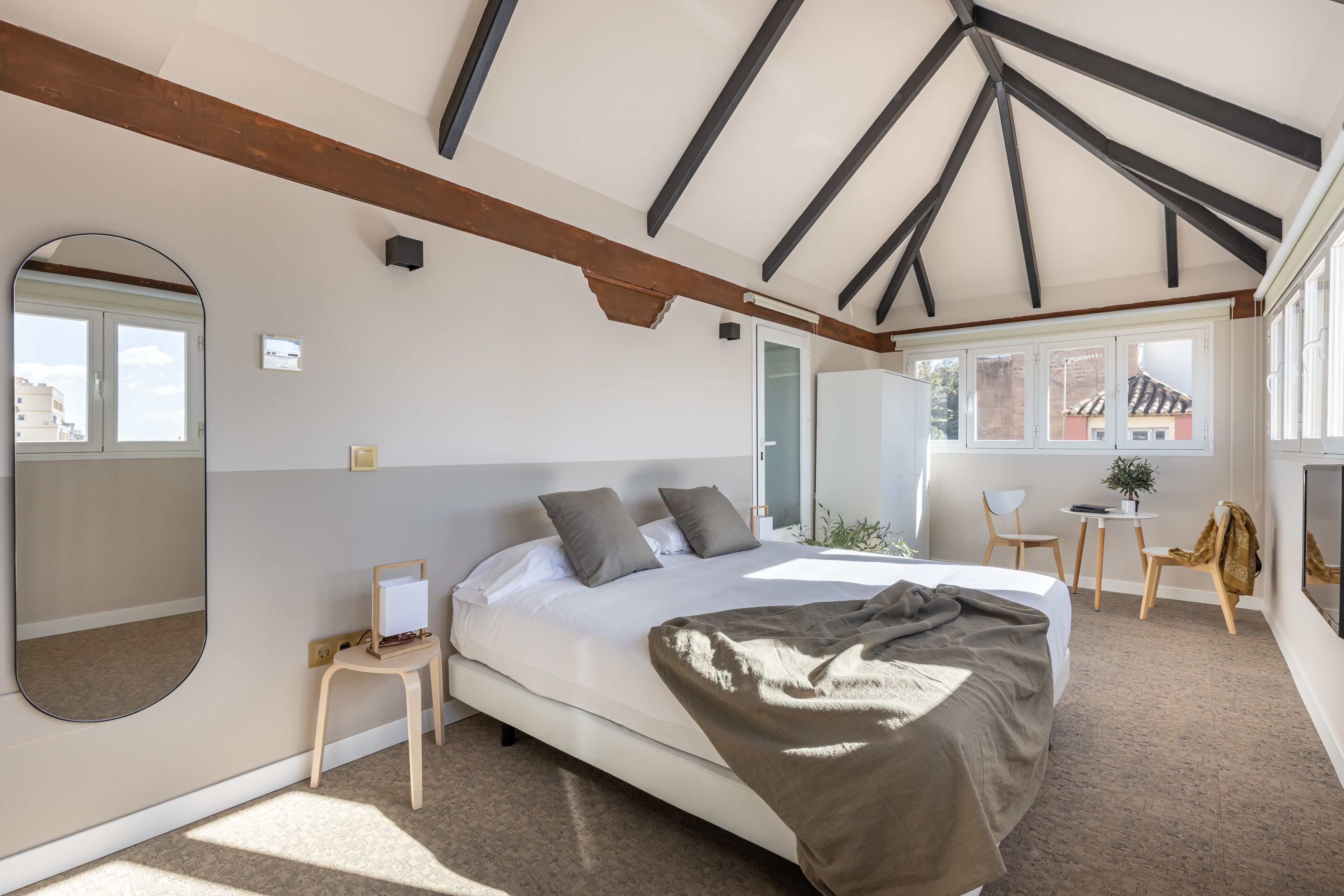 Un dormitorio moderno con una cama grande, un escritorio pequeño, una silla, un espejo y una ventana con vistas a edificios.