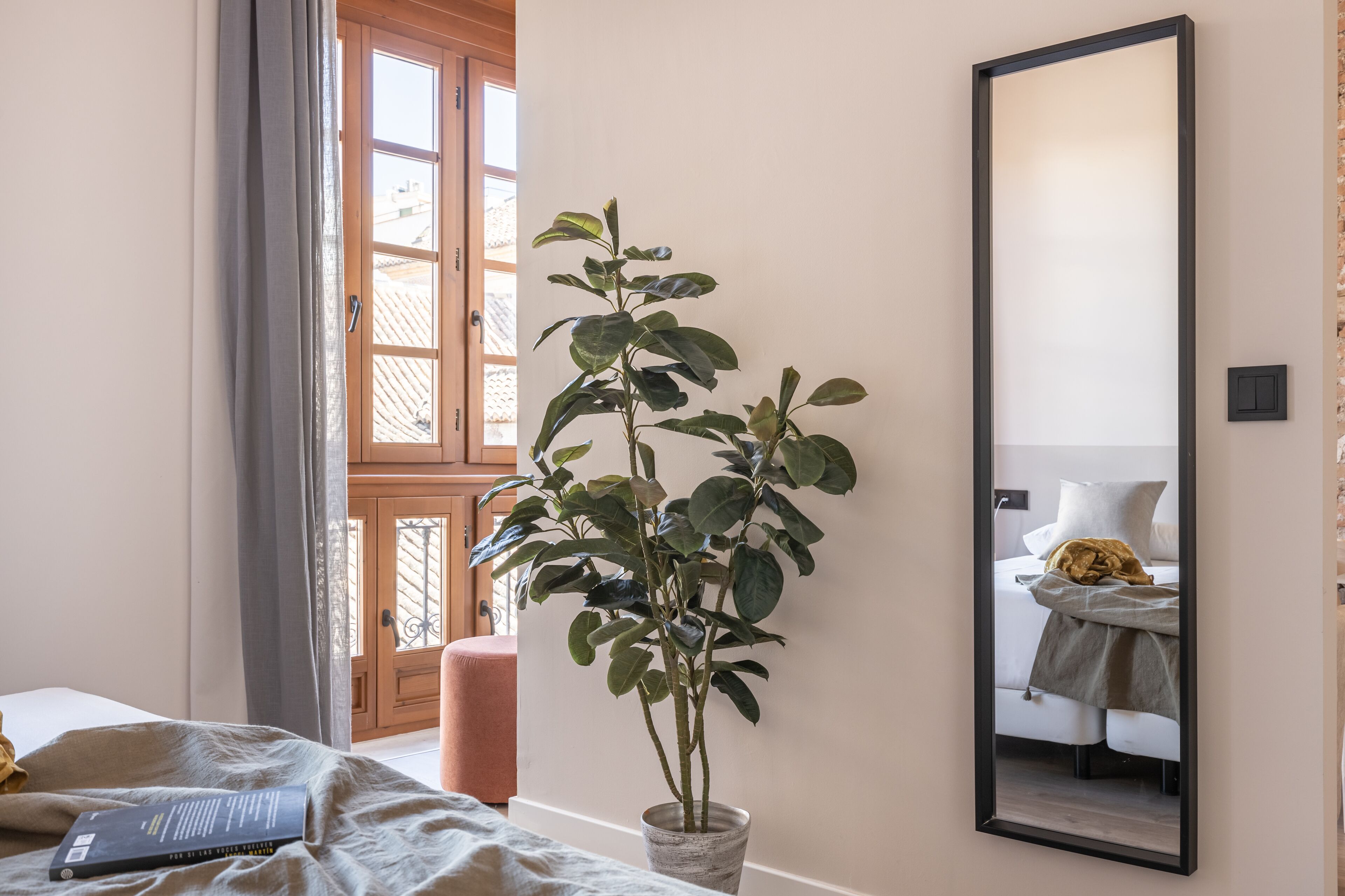 A bedroom with a potted plant, a bed, a bedside table with a book, a mirror, and a window with curtains.