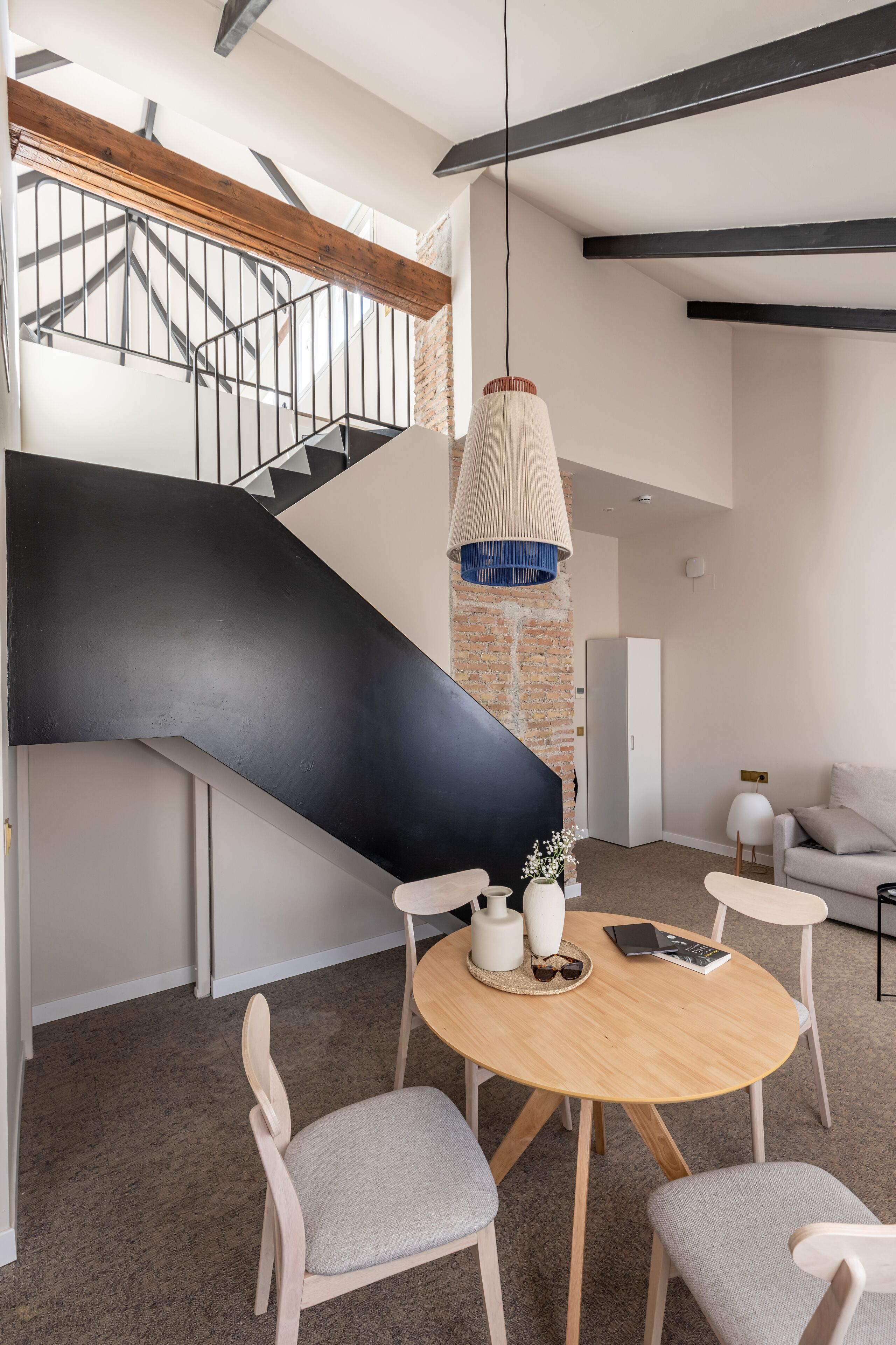 A modern interior with a wooden dining table, chairs, a staircase with a black accent, and a hanging light fixture.
