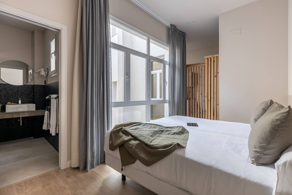 Een slaapkamer met een bed, een raam met gordijnen, een houten garderobe en een badkamer met een spiegel en wastafel.