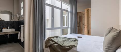 Un dormitorio con cama, ventana con cortinas, armario de madera y baño con espejo y lavamanos.
