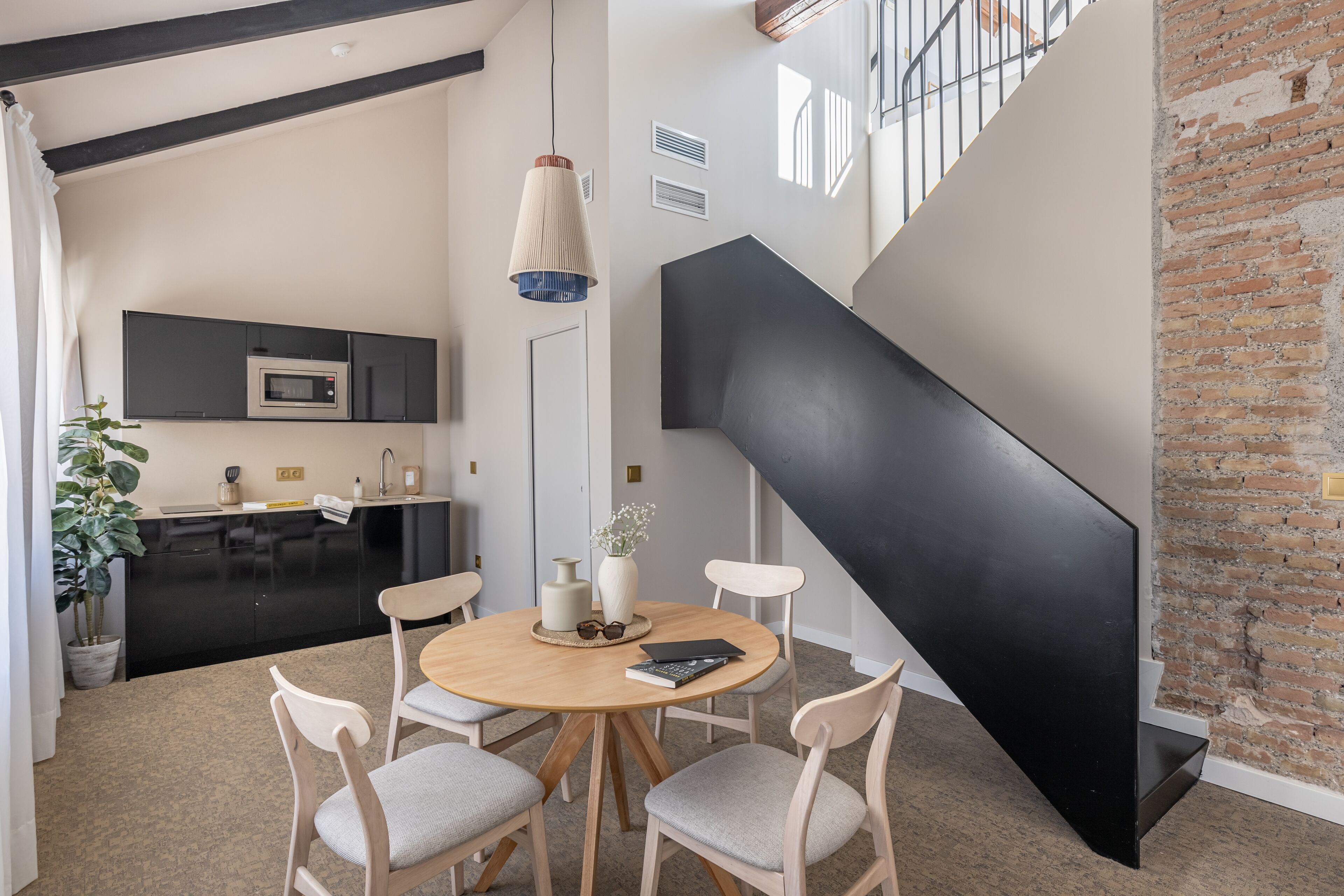 Una cocina moderna con armarios negros, un microondas y un lavabo. Hay una zona de comedor con una mesa redonda y cuatro sillas. Una escalera con barandilla negra lleva a un nivel superior.