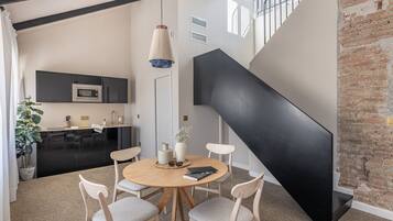 Una cocina moderna con armarios negros, un microondas y un lavabo. Hay una zona de comedor con una mesa redonda y cuatro sillas. Una escalera con barandilla negra lleva a un nivel superior.