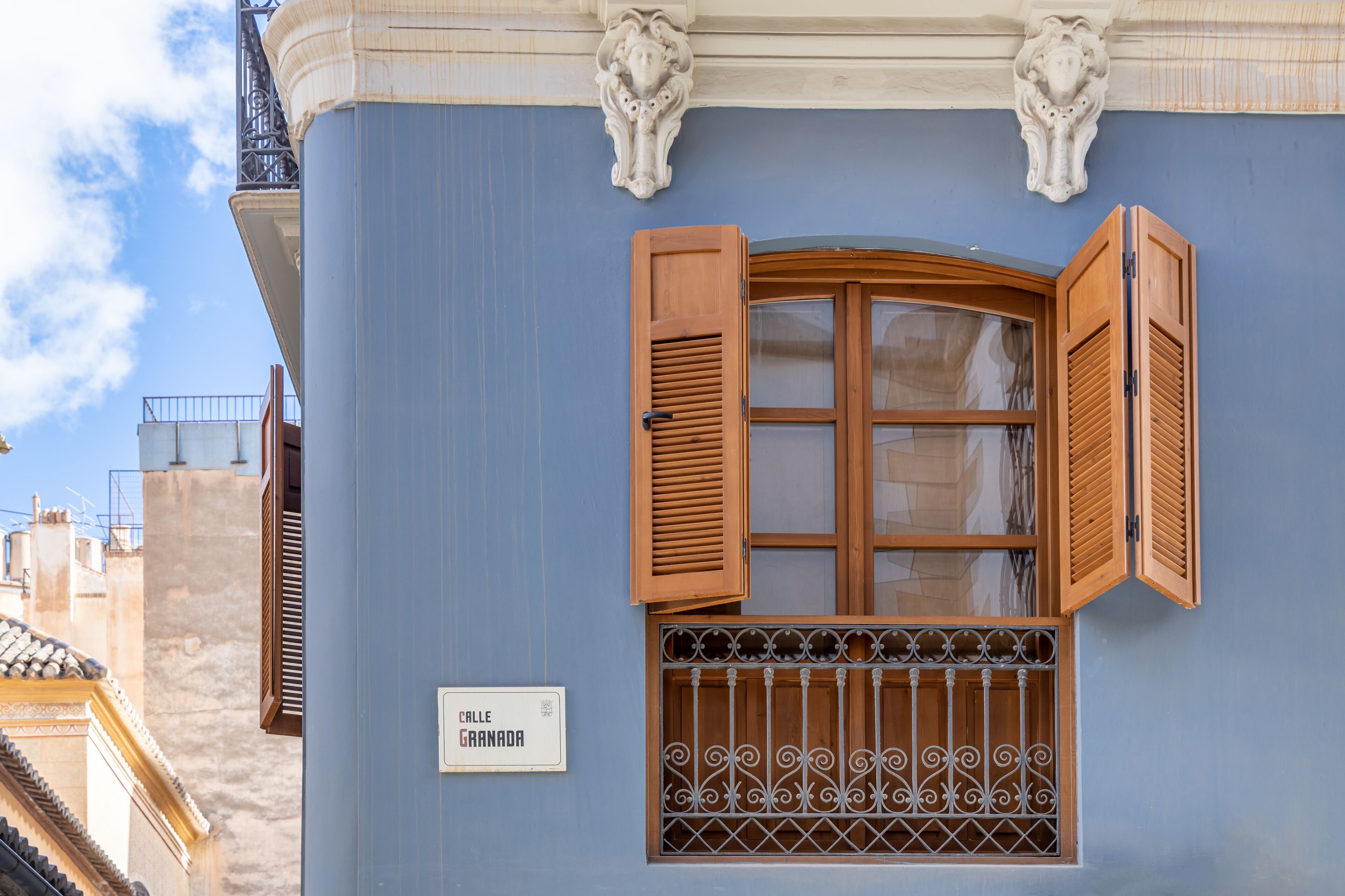 A building facade with a blue exterior, a wooden window with shutters, a metal grille, and a sign reading "Calle Granada".