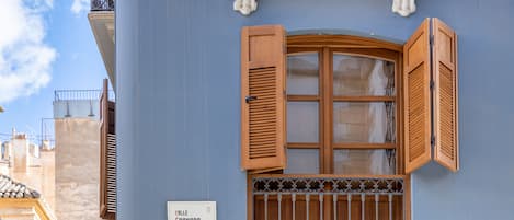 Facciata di un edificio con esterno blu, una finestra in legno con persiane, una griglia metallica e un cartello con la scritta