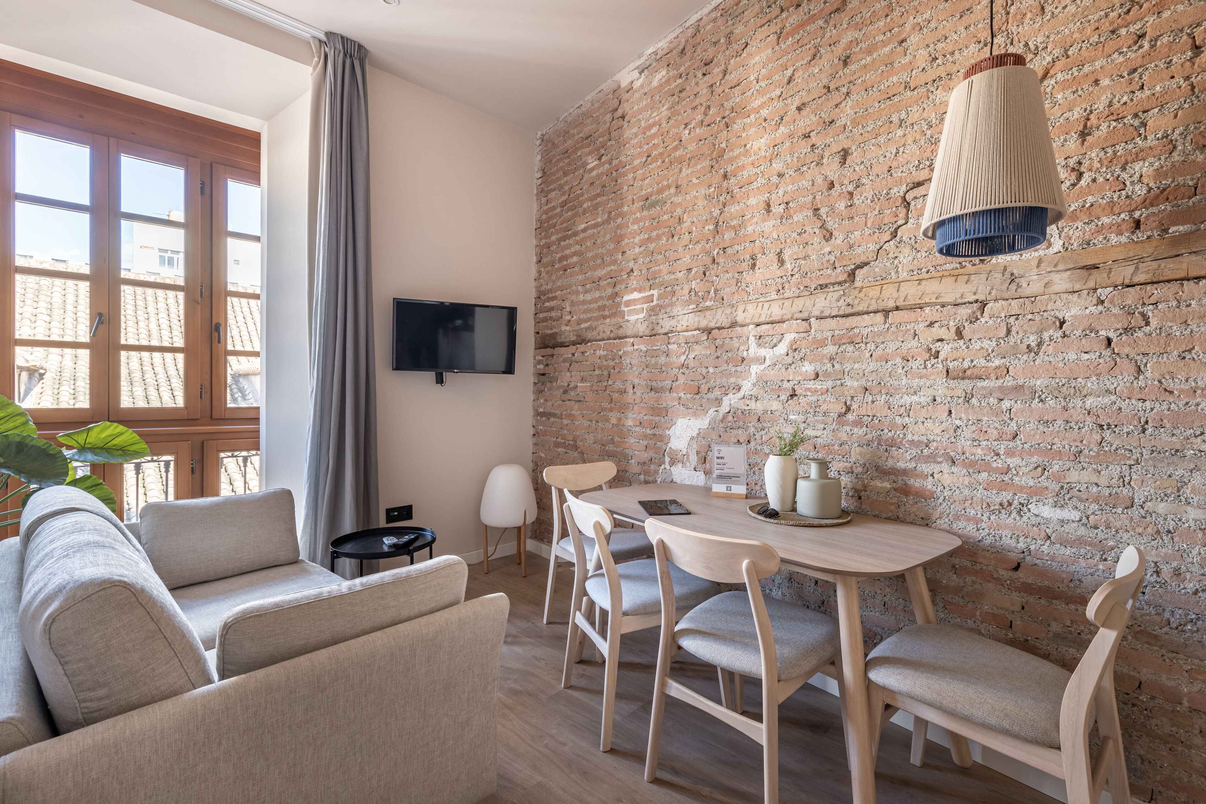 A modern living room with a brick wall, a flat-screen TV, a dining table with chairs, and a sofa.