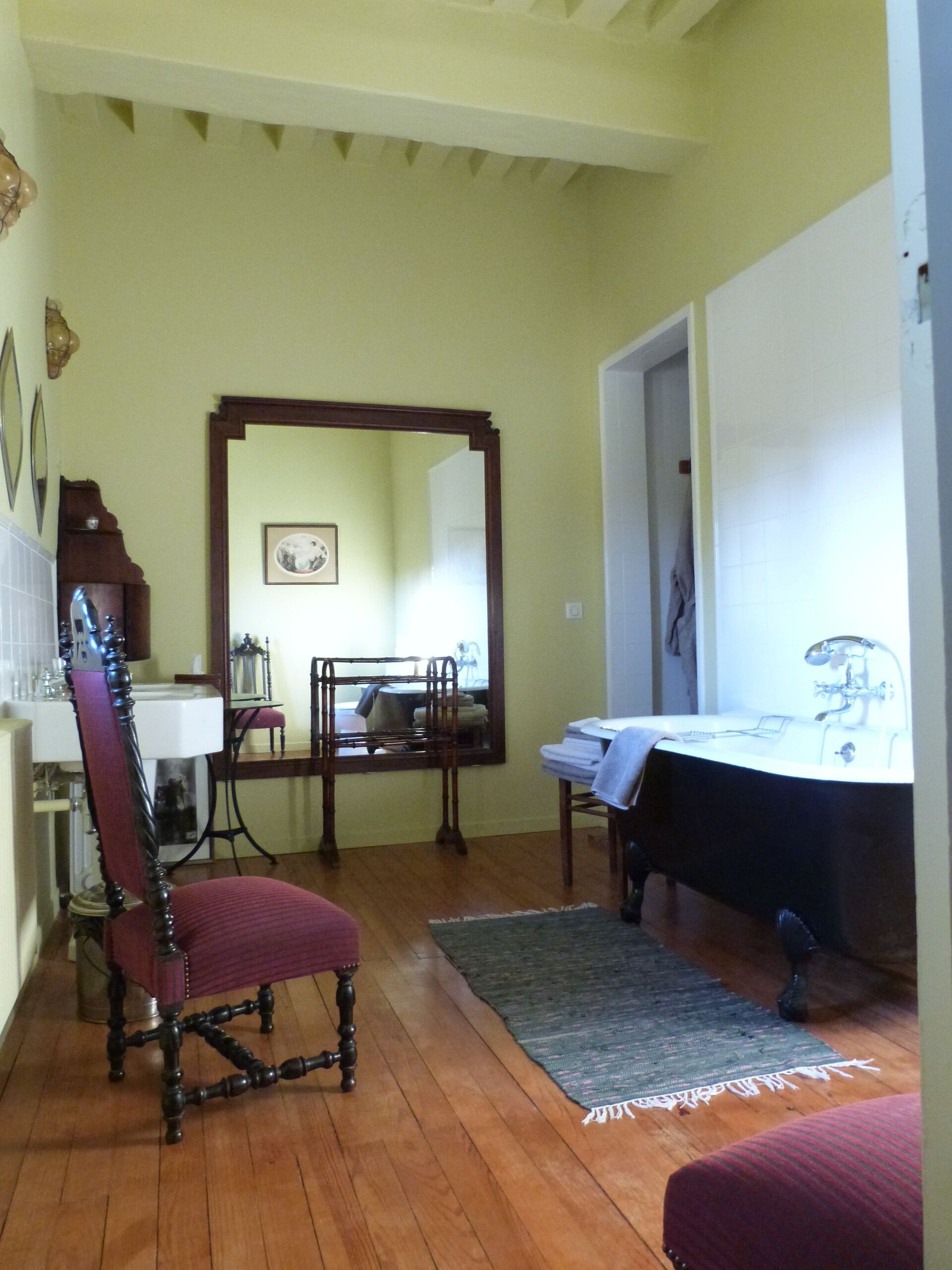 Une pièce comprenant une chaise en bois, un lit recouvert d’une couverture blanche, un miroir et une petite table avec une lampe.