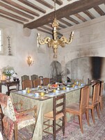Uma sala de jantar com uma mesa comprida posta para refeição, rodeada por cadeiras de madeira. Um lustre pendurado no teto, e uma lareira visível ao fundo.