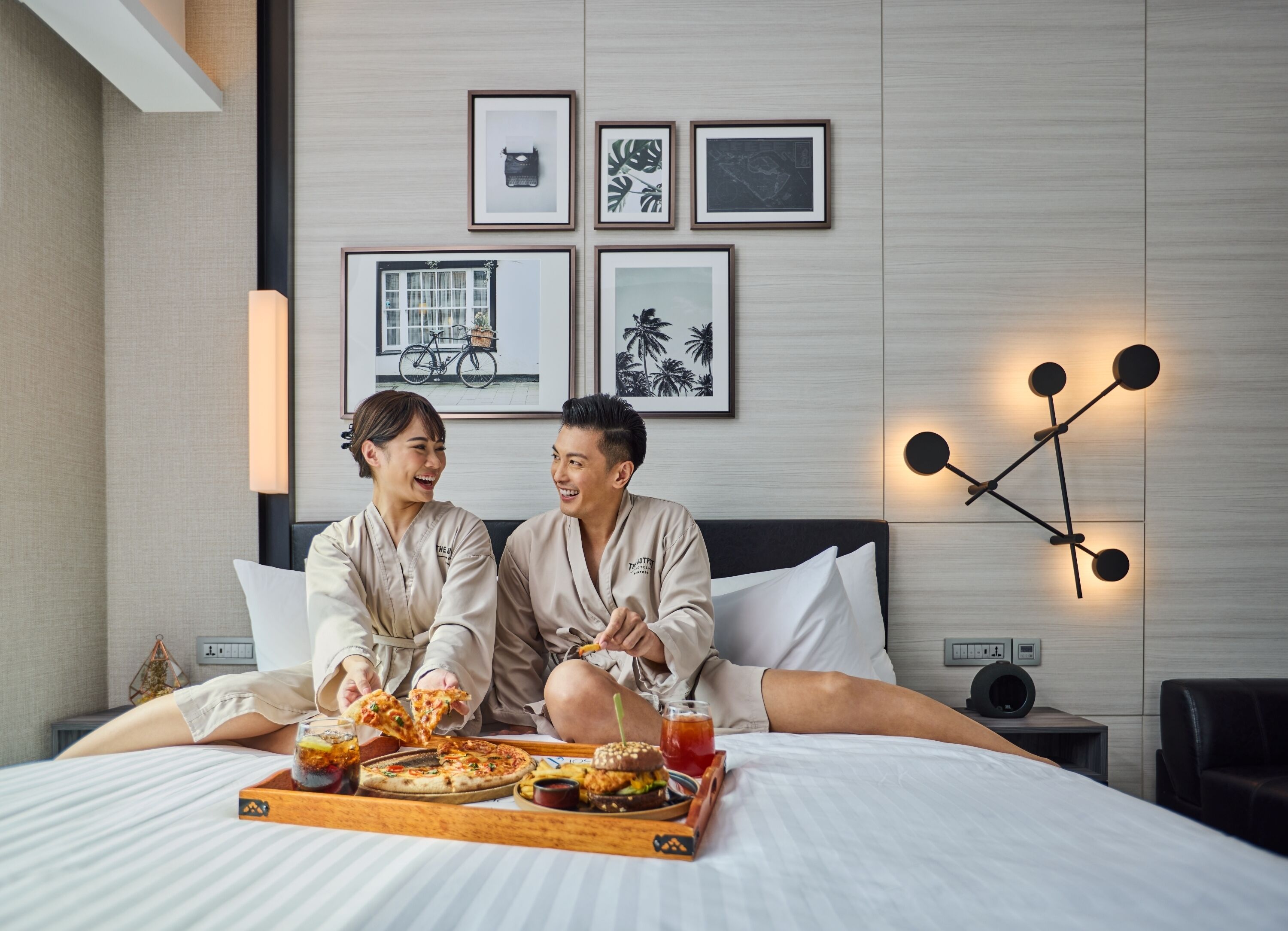 A hotel room with a bed, a tray with pizza and drinks, framed pictures on the wall, and a modern light fixture.