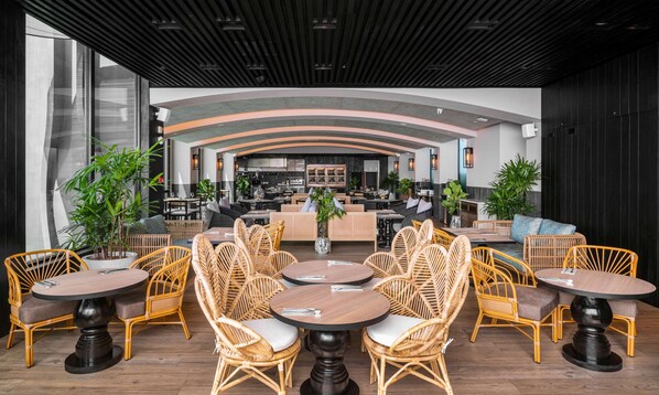 A modern restaurant with round tables and wicker chairs, a bar area, and a view of the cityscape.
