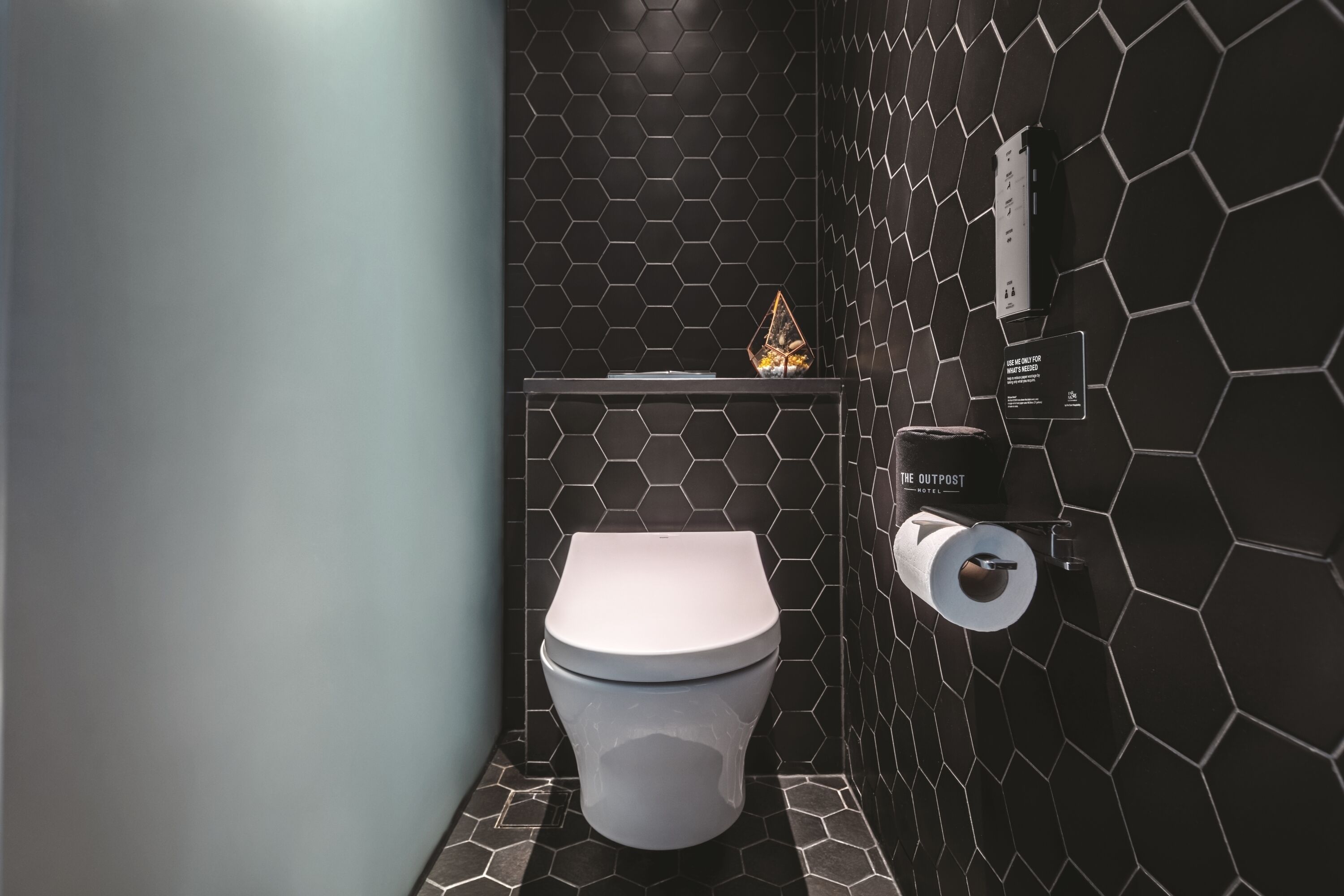 A modern bathroom with a hexagonal tile pattern, a toilet, a trash bin, and a soap dispenser on the wall.