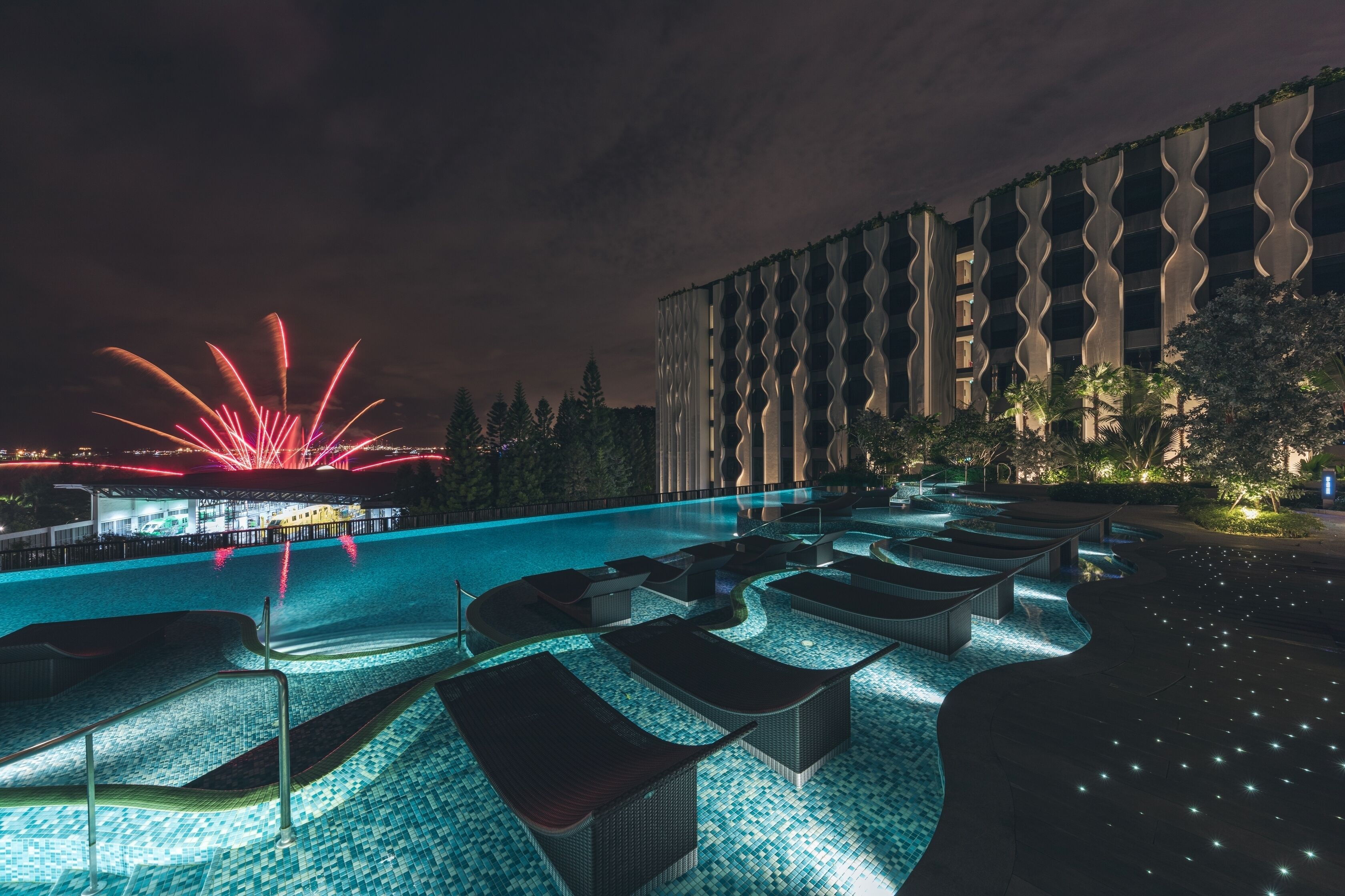 A nighttime pool area with lounge chairs and a building with a unique wavy facade.