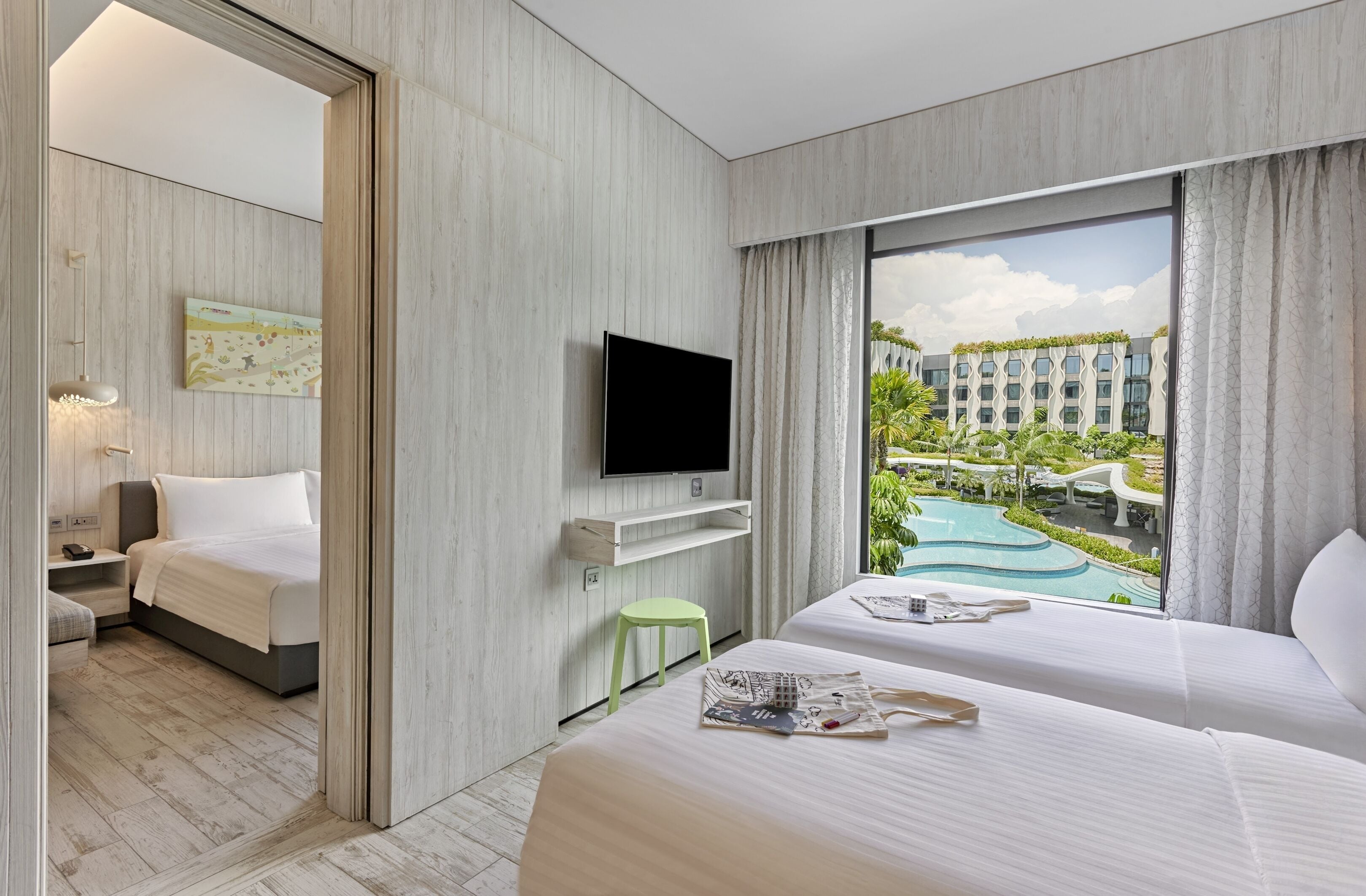 A hotel room with a bed, a TV mounted on the wall, a small stool, and a window with a view of a pool and buildings.