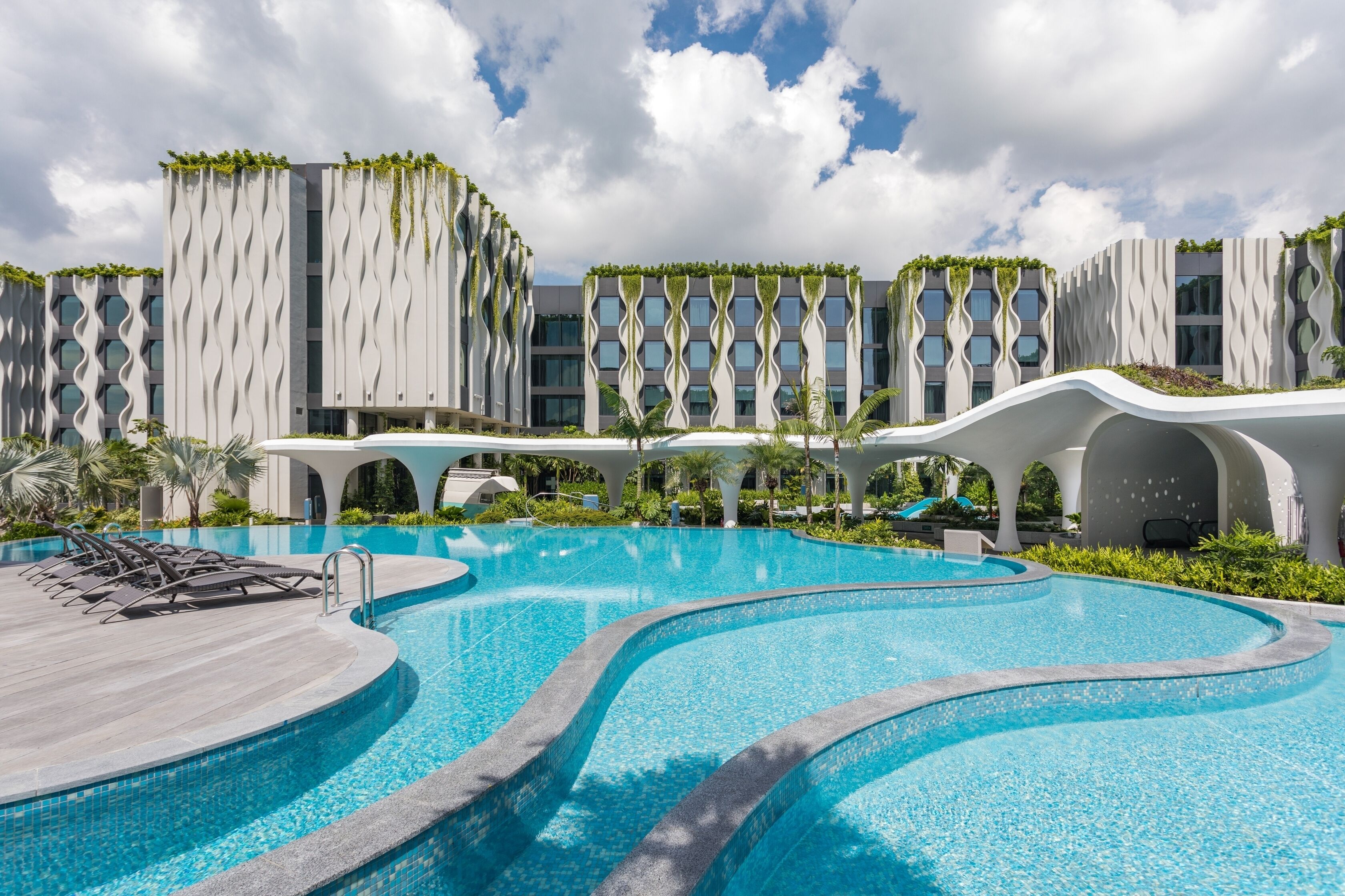 A modern hotel with a large swimming pool, unique architecture, and lush greenery.