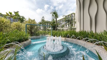 A pool with multiple water jets surrounded by tropical plants and a modern building with unique architecture in the background.