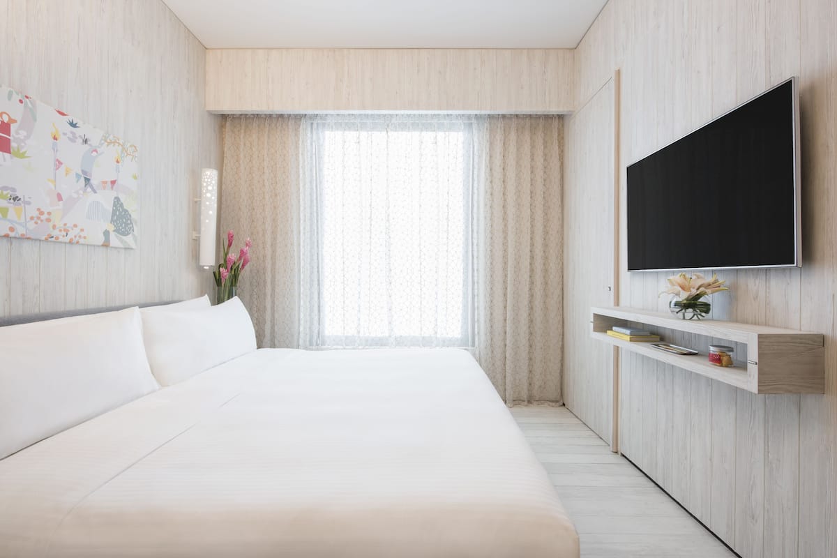 Une chambre d’hôtel moderne dotée d’un grand lit, d’un téléviseur à écran plat fixé au mur et d’une fenêtre avec des rideaux légers.