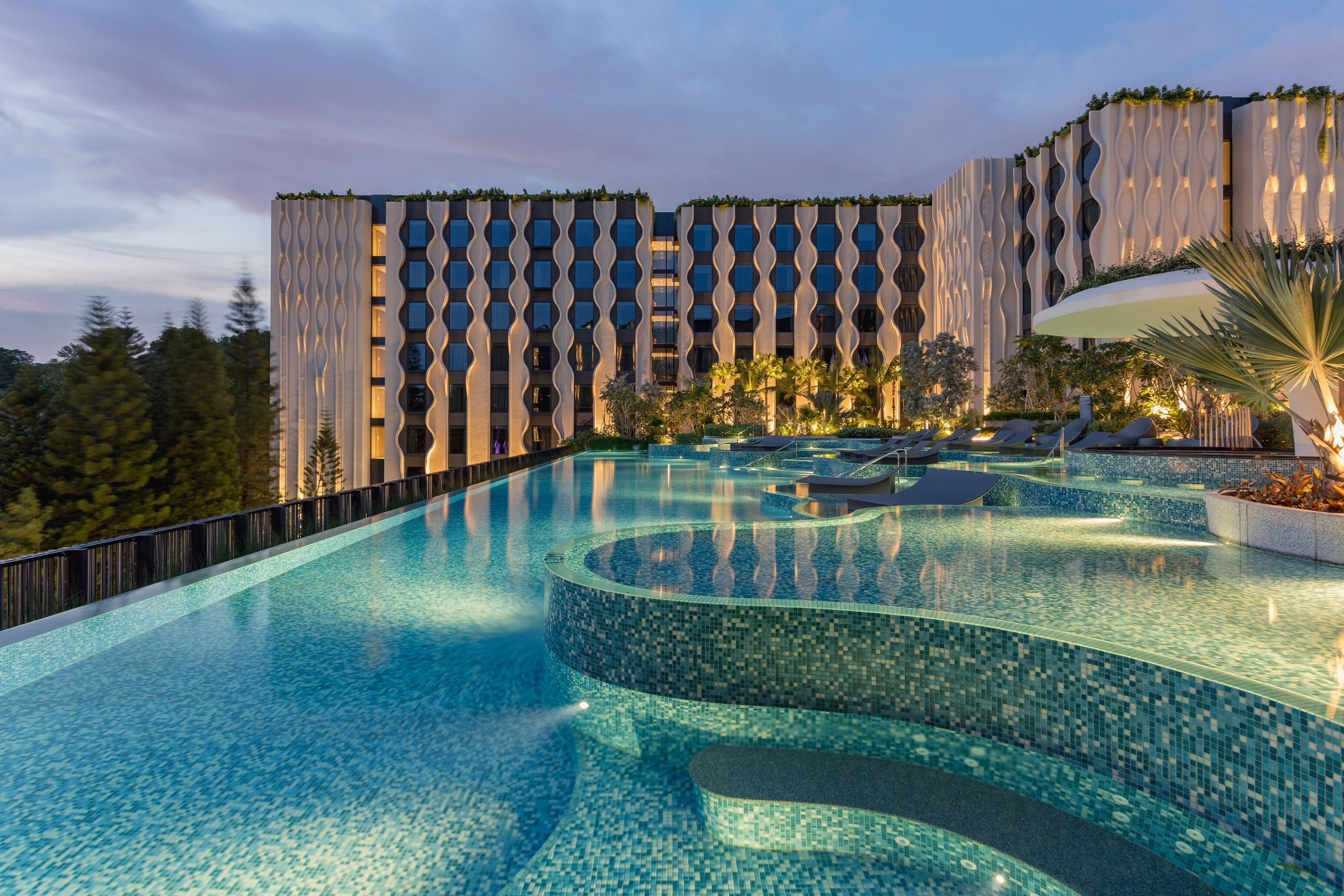 A modern hotel with a large infinity pool, a wavy exterior wall, and a well-maintained garden.