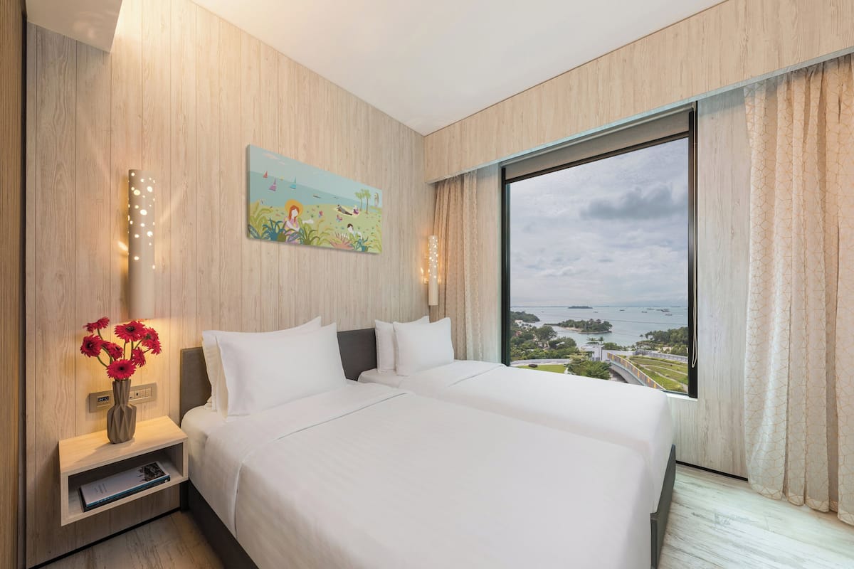 Une chambre d’hôtel avec un grand lit, une table de chevet ornée d’un vase de fleurs, un tableau accroché au mur et une vue sur la mer par une grande fenêtre.