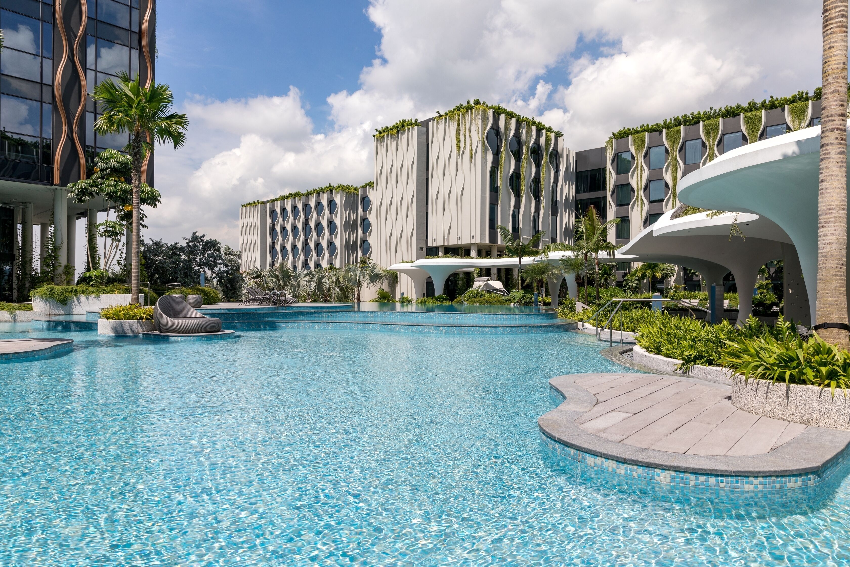 Un hotel moderno con una piscina centrale, circondato da palme e una facciata ondulata.