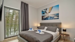 A modern hotel room with a large bed, a window with curtains, a painting of a boat on the sea, and a small table with a lamp. - Willa Tamara BlueApart Jurata (Jurata)