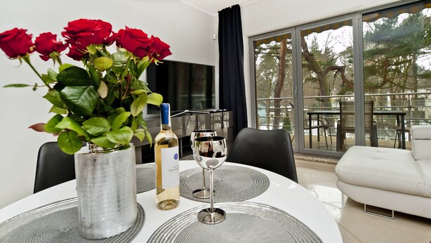 A modern dining area with a round table, a vase of red roses, a bottle of wine, and a wine glass. - Willa Tamara BlueApart Jurata (Jurata)