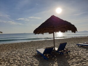 Una spiaggia con due sdraio blu sotto un ombrellone di paglia, sabbia fine e l'oceano sullo sfondo.