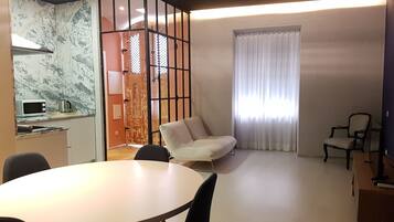Una sala de estar moderna con una mesa de comedor redonda, un sofá y una silla. Hay una zona de cocina con microondas y hervidor.