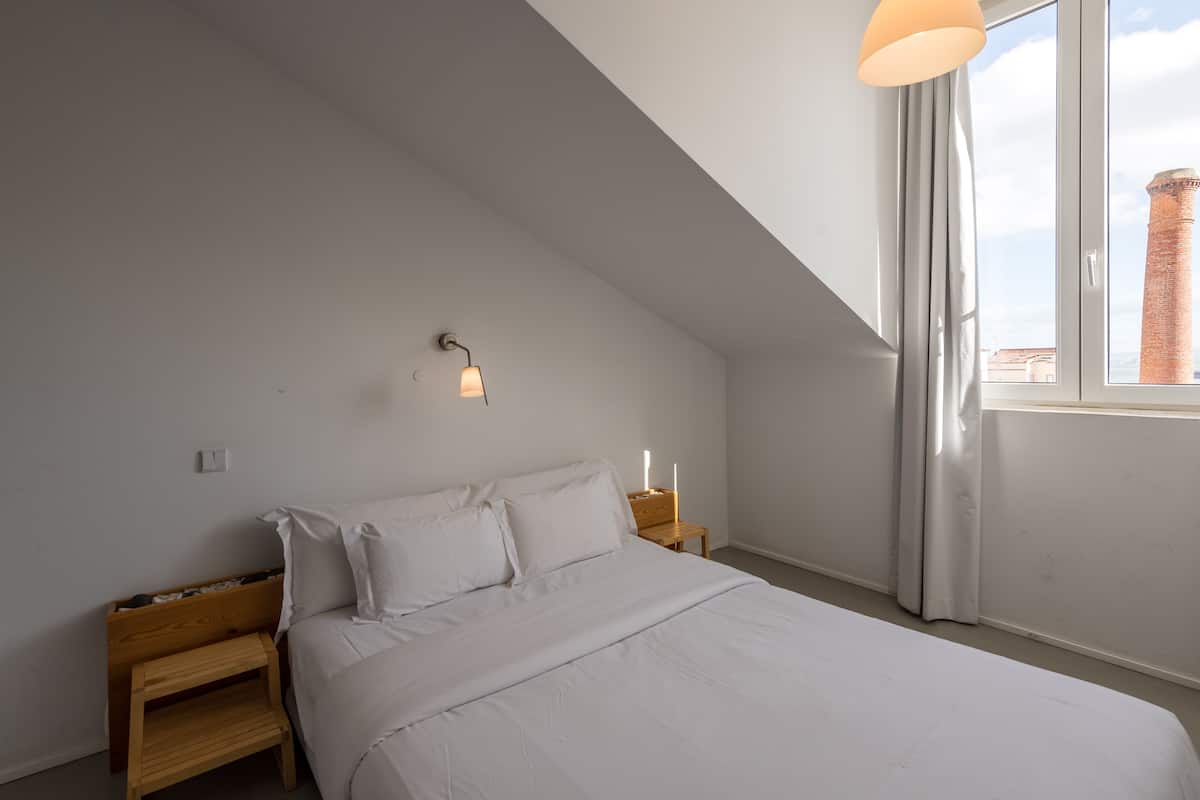 Un dormitorio con techo inclinado, una cama con ropa blanca, una mesita de noche con lámpara y vistas a una torre de ladrillo a través de una ventana.