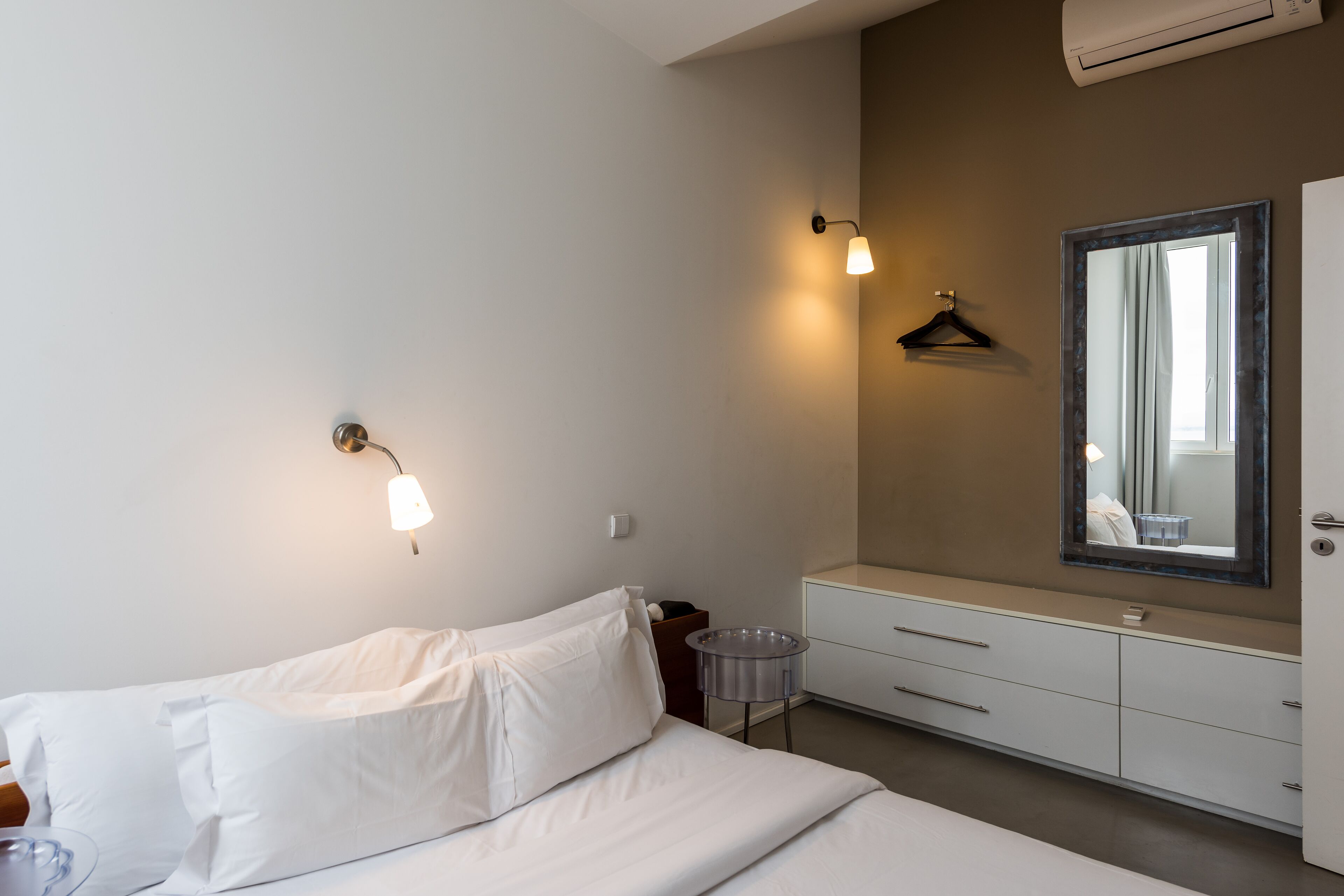 A hotel room with a bed, bedside table, dresser, mirror, and wall-mounted light fixture.