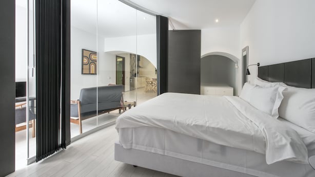 Un dormitorio moderno con una cama grande, un ventanal de vidrio que da a un balcón y vistas al salón.