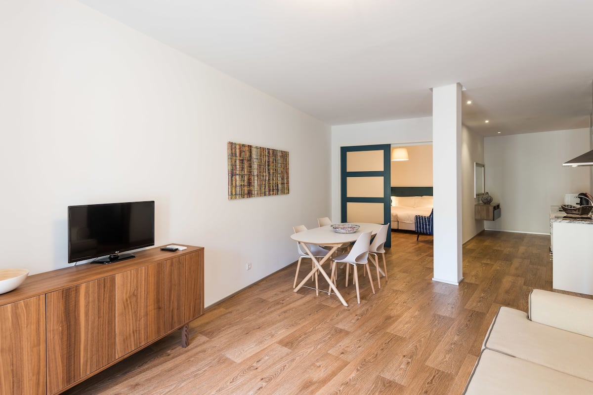 Una sala de estar moderna con un televisor de pantalla plana sobre un mueble de madera, un comedor con una mesa redonda y sillas, y una zona de cocina con estufa y armarios.