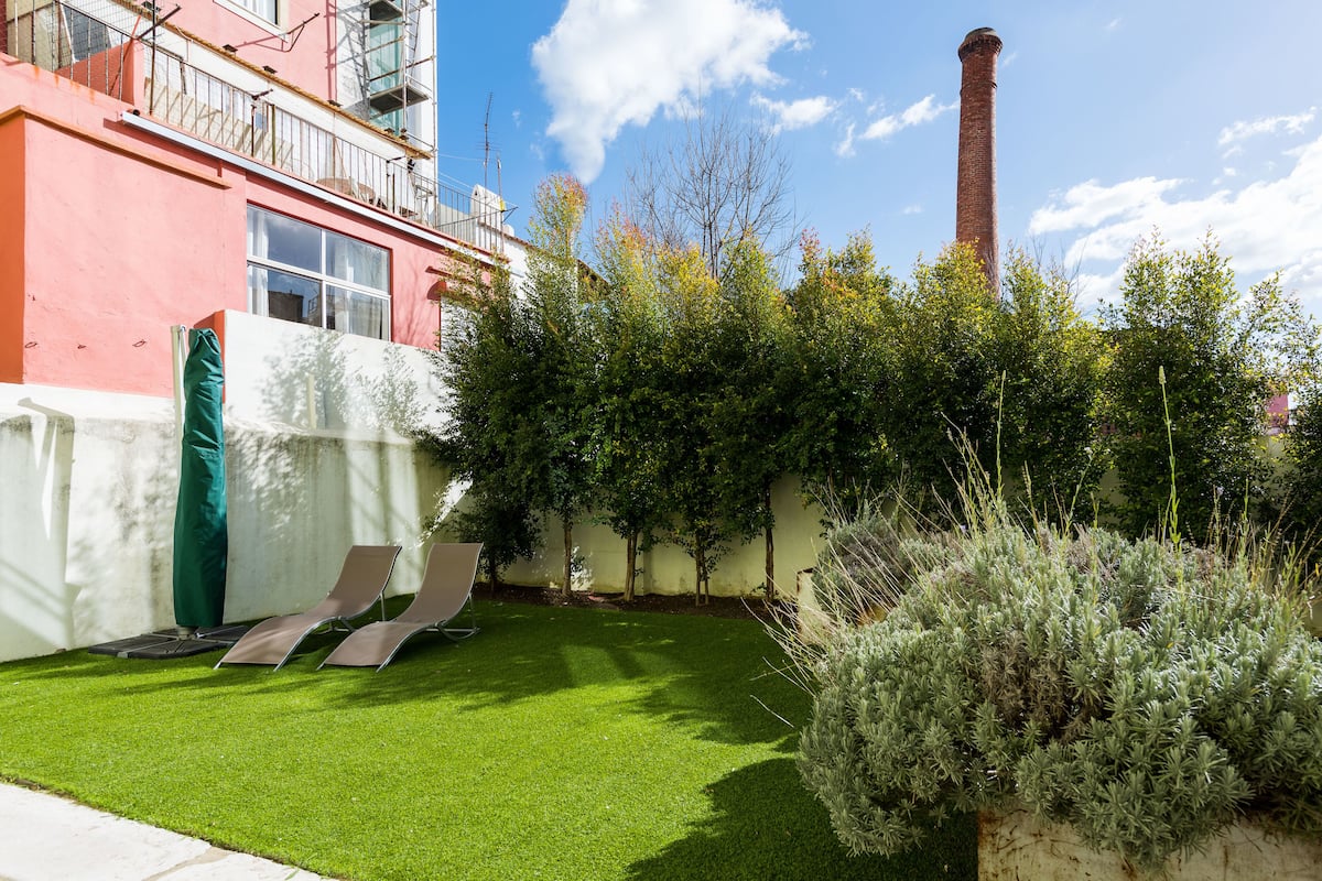 Un patio con césped artificial, dos sillones de descanso, un parasol verde y una chimenea alta al fondo.