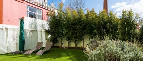 A courtyard with artificial grass, two lounge chairs, a green umbrella, and a tall chimney in the background.