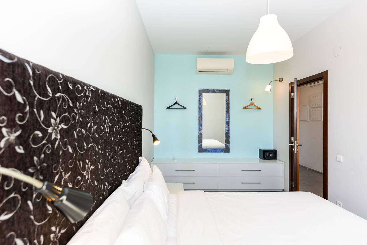 Un dormitorio con cama, lámpara de noche, cómoda, espejo y lámpara colgante.