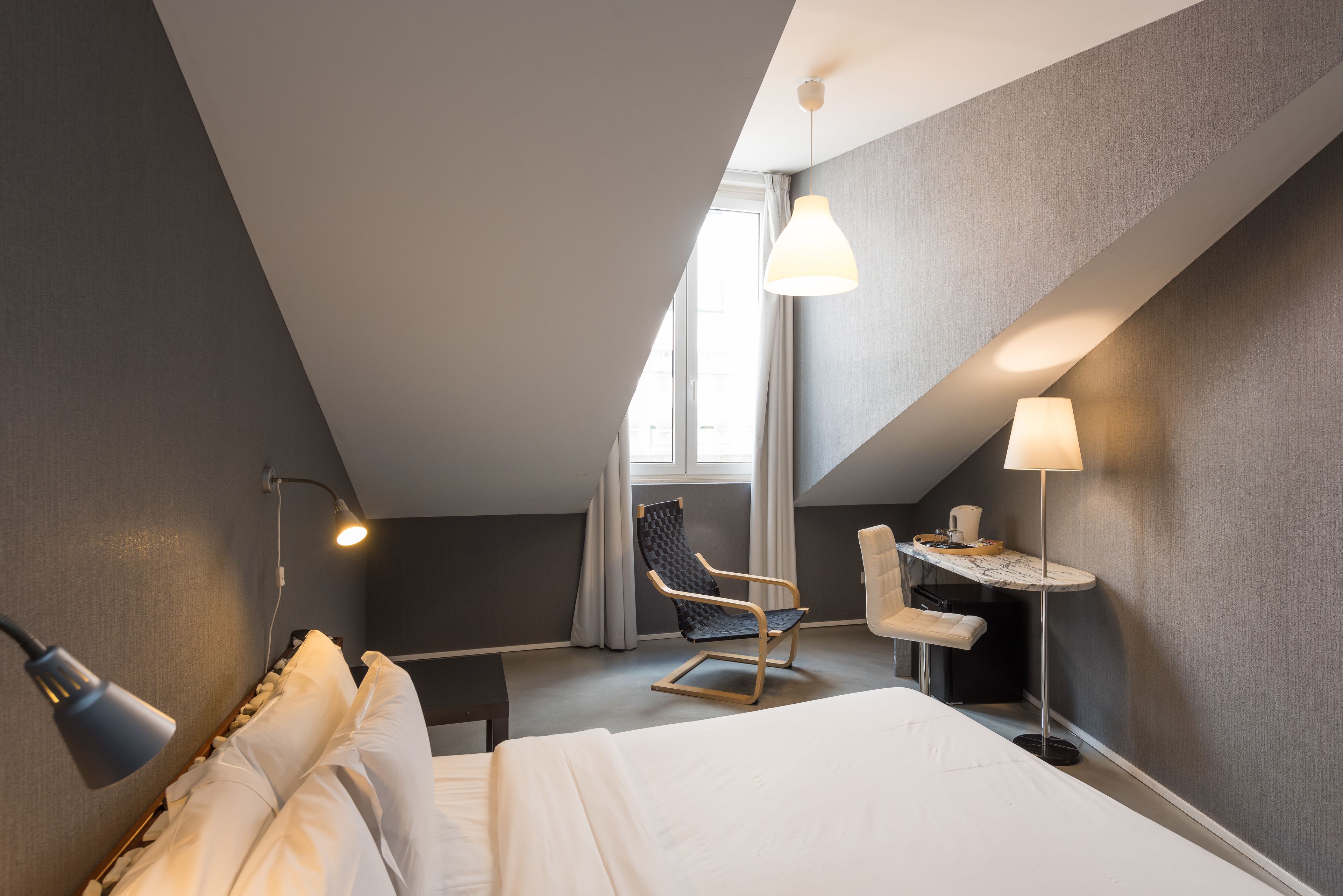 A modern hotel room with a large bed, a desk with a lamp, a chair, and a window with curtains.