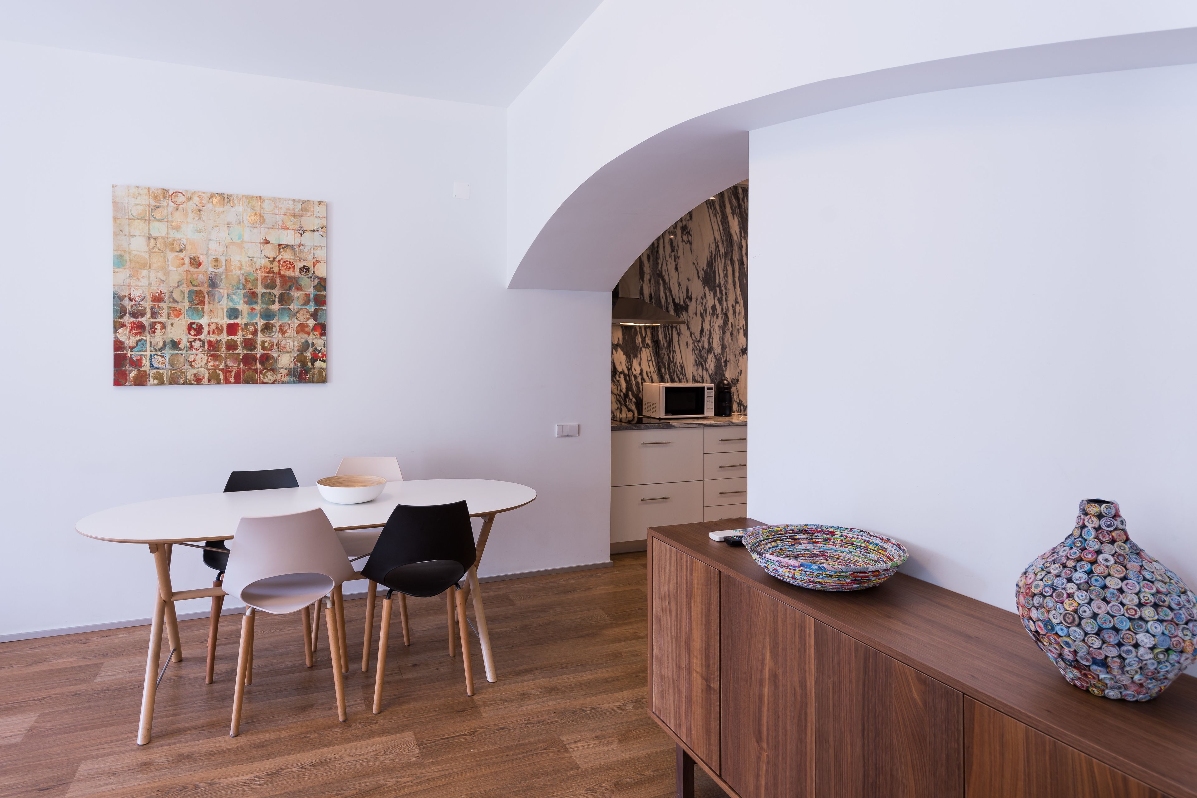 Uma zona de refeições moderna com uma mesa e cadeiras brancas, um armário de madeira com objetos decorativos e uma cozinha ao fundo.