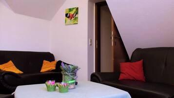 Un salon avec deux canapés noirs, dont l’un est agrémenté de coussins orange et l’autre de coussins rouges, une petite table sur laquelle est posé un vase de fleurs, et un tableau représentant un papillon accroché au mur.