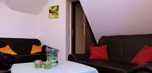 A living room with two black sofas, one with orange and one with red cushions, a small table with a vase of flowers, and a butterfly painting on the wall. - Landgasthaus Veltheimer Hof (Porta Westfalica)