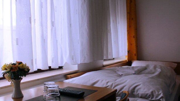 A hotel room with a bed, a bedside table with a vase of flowers, a remote control, and a window with white curtains. - Landgasthaus Veltheimer Hof (Porta Westfalica)