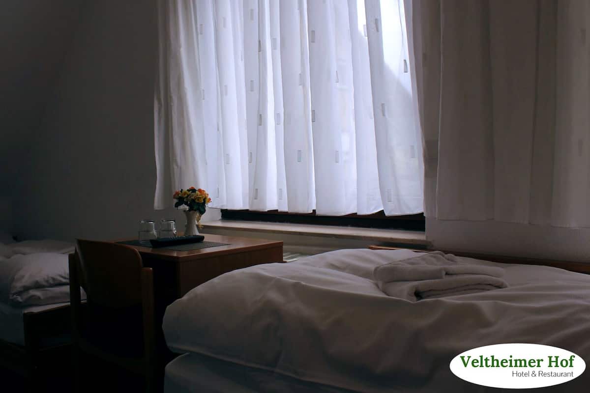 A hotel room with two beds, a small table with a vase of flowers, and a window with sheer curtains.