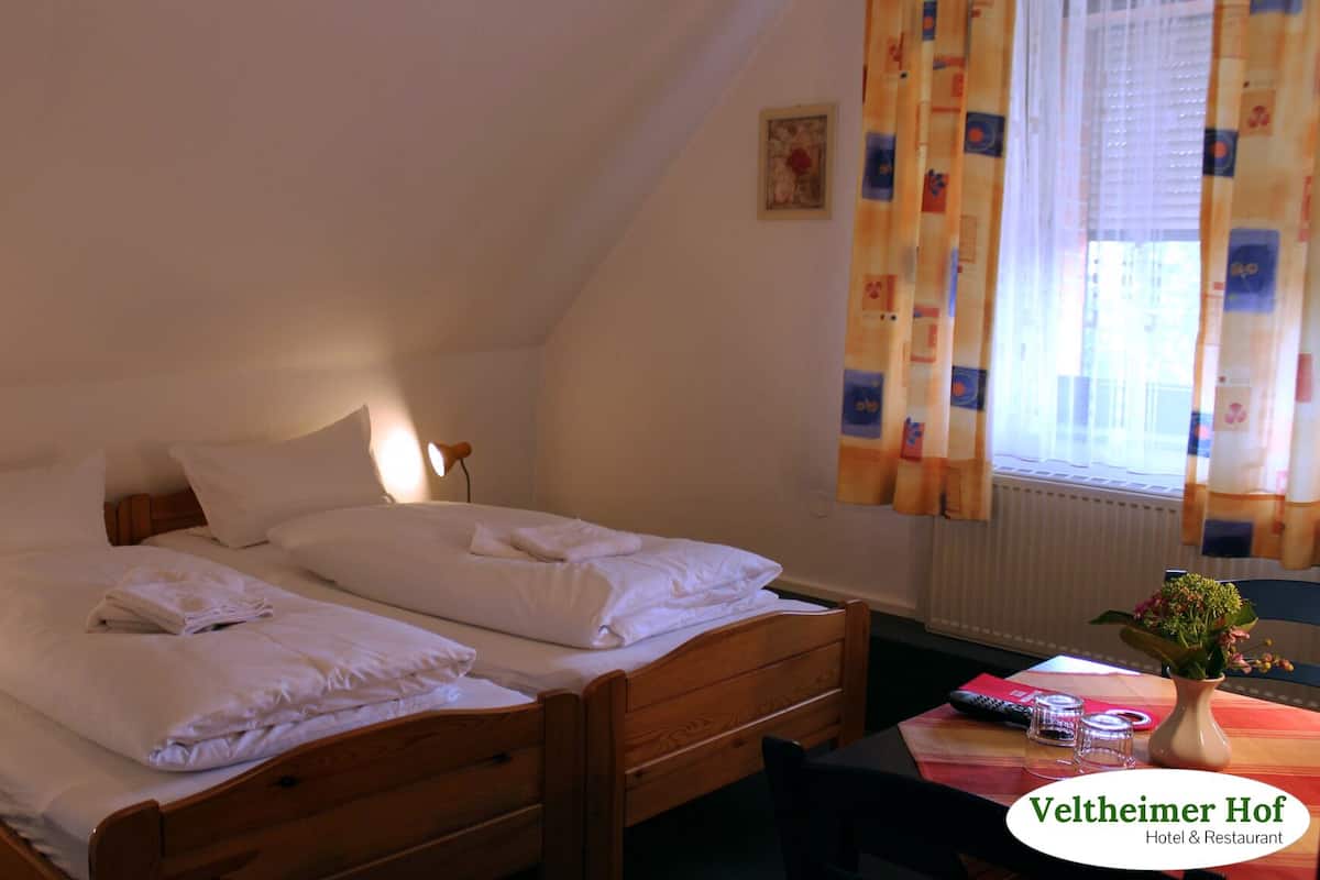 A room with two wooden beds, a small table with a vase and remote control, and a window with curtains.