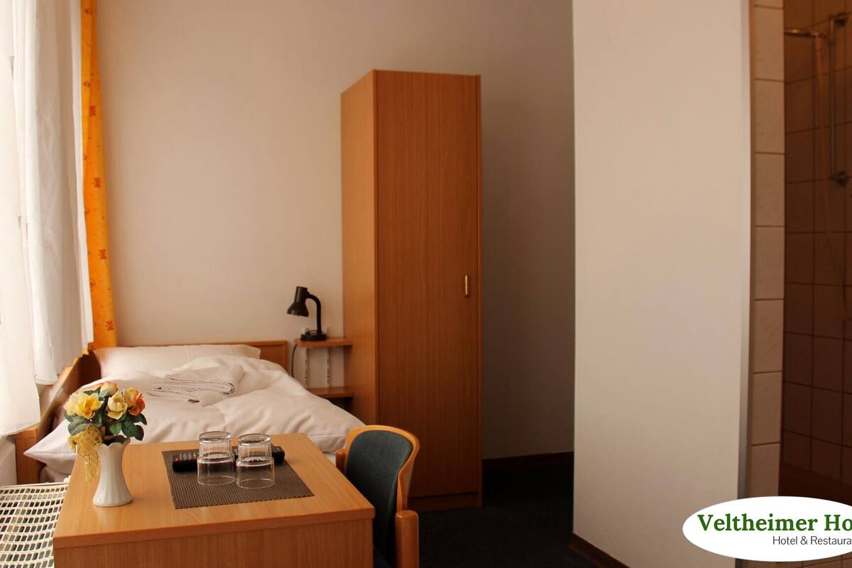 A hotel room with a bed, a desk with a vase of flowers, a lamp, and a wardrobe.