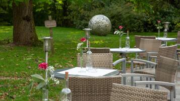 Buiten zitplaatsen met rieten stoelen en tafels, omringd door groen en een grote decoratieve bol.