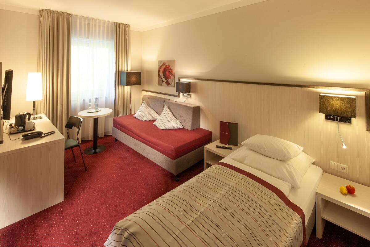 Habitación de hotel con cama, escritorio, silla, mesita, lámpara, cuadro y mesita de noche con lámpara.