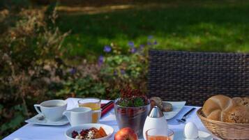 An outdoor dining table with breakfast items, including eggs, bread, and fruit, set in a garden with trees and flowers in the background.