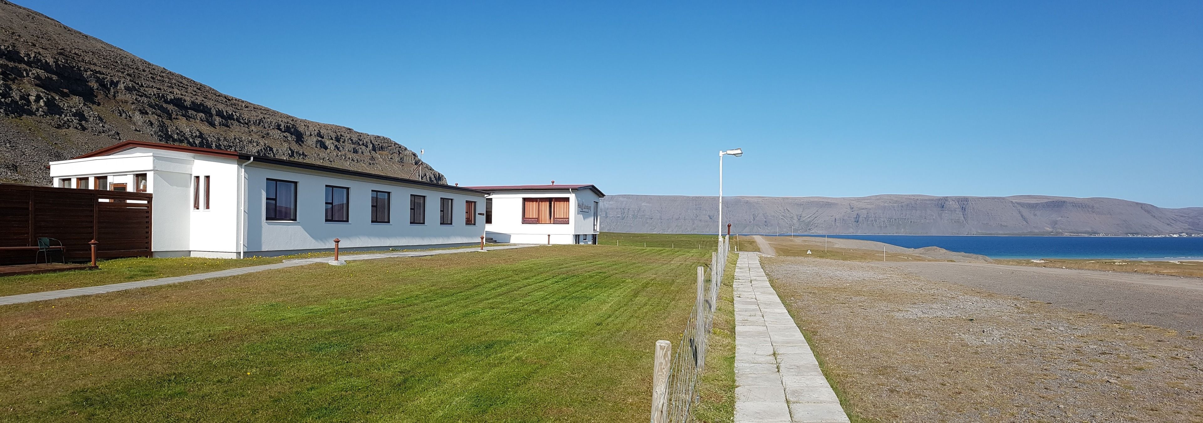 A white building with a red roof, a paved pathway, and a view of the sea and mountains.