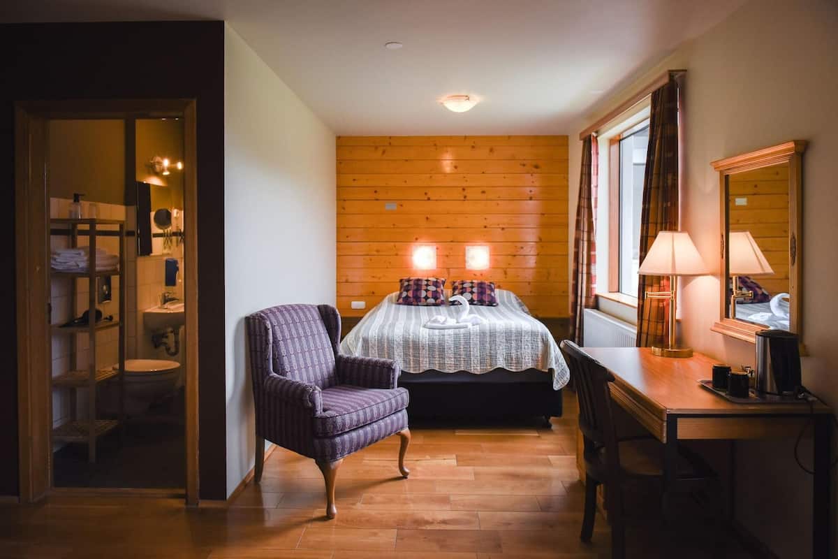 Un dormitorio con cabecera de madera, cama con ropa de cama estampada, una silla, un escritorio con lámpara y un espejo.