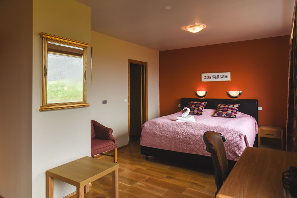 Un dormitorio con una cama, una silla de madera, una mesita, una ventana y una lámpara fijada a la pared.