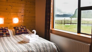 Habitación de hotel con pared de madera, un ventanal con vistas a un paisaje cubierto de hierba y montañas, una cama con ropa de cama estampada, y dos lámparas fijadas a la pared.